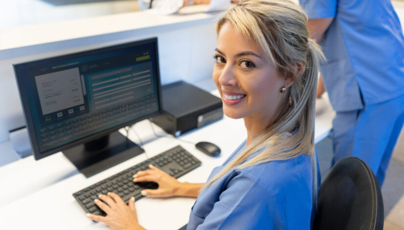 Beautiful friendly nurse at the front desk of the hospital working on computer while facing camera smiling
