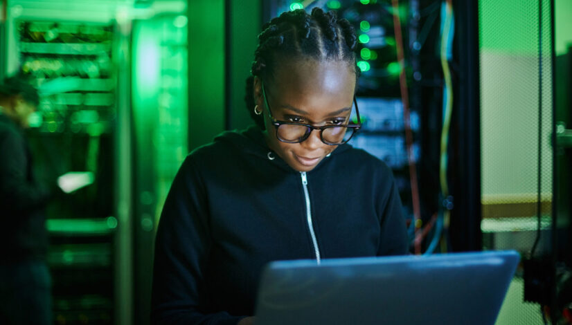 Shot of a young female computer programer using a laptop while standing in a dark server room